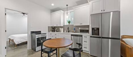 Une cuisine compacte avec des meubles blancs, des appareils en acier inoxydable, une table à manger avec des chaises et un lit dans la pièce adjacente.