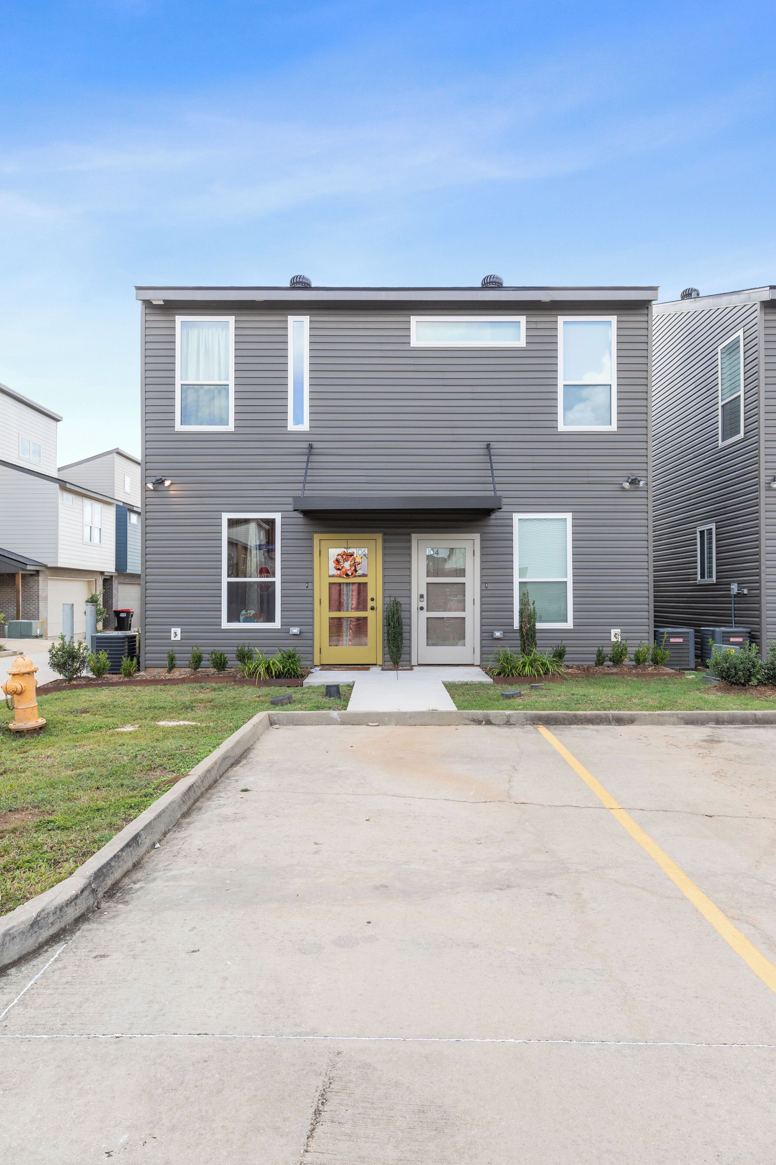 A two-story residential building with a front entrance, a small front yard with plants, and a parking area in front.