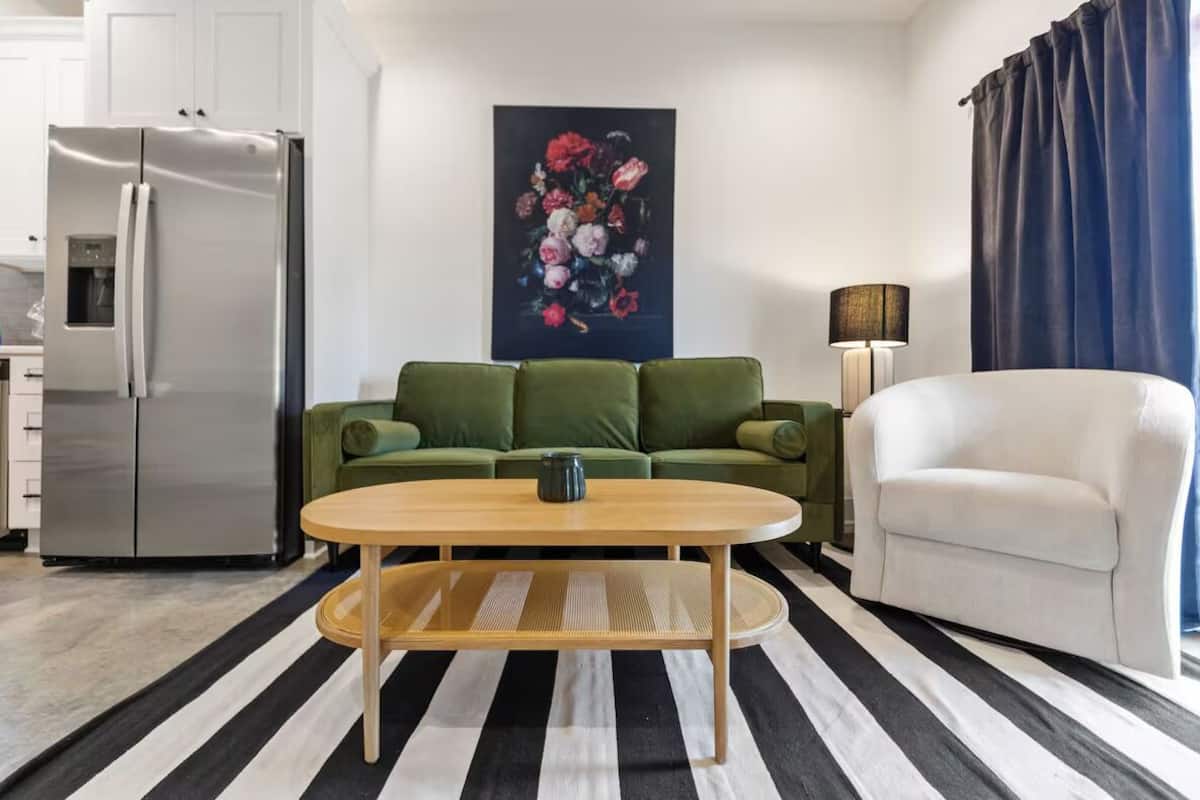 A modern living room with a striped rug, a green sofa, a white armchair, and a painting of flowers on the wall.