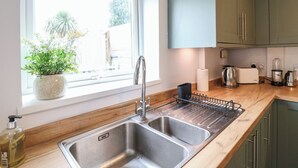 Une cuisine équipée d’un évier à deux bacs, d’une fenêtre donnant sur un espace verdoyant et d’un plan de travail sur lequel se trouvent une bouilloire et un grille-pain.