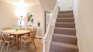Un espace repas avec une table et des chaises en bois, un escalier recouvert d’une moquette à motifs et un luminaire au plafond.