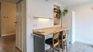 Une cuisine compacte avec un plan de travail en bois, deux chaises, une étagère intégrée ornée de plantes et un réfrigérateur blanc.