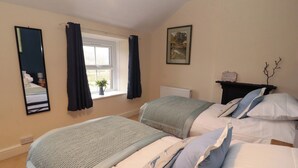 A bedroom with two beds, a window with curtains, a radiator, a framed picture, and a small plant on the windowsill. - Easthwaite Cottage (Seascale)