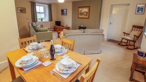A dining area with a wooden table set for four, surrounded by chairs. There is a sofa and a rocking chair in the background. - Easthwaite Cottage (Seascale)