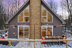 A two-story house with a central brick chimney, large windows, and a deck with outdoor seating. - A-Frame Escape ~ Hot Tub, Sauna, Game Room, Gym (Big Bass Lake)