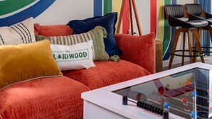 A vibrant room with a neon sign reading "Lodge and Play" on the wall, a red sofa with multiple cushions, a floor lamp, and a glass coffee table with a boxing ring miniature.