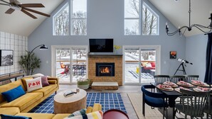 A spacious living room with a fireplace, large windows, a ceiling fan, and a dining area with a table set for a meal.