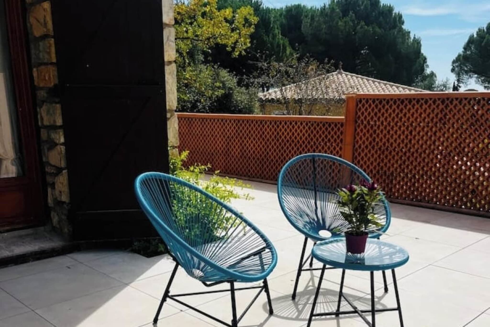 A patio with two blue chairs and a small table, surrounded by a wooden fence and greenery.