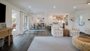 A spacious living area with a wooden dining table, a sofa, and a kitchen in the background. - 22 Sand Dollar - Koi Cottage: Island Serenity, 1st Row, Folly Field (Hilton Head Island)