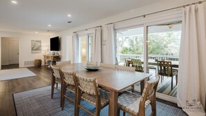 A spacious dining area with a wooden table and chairs, large windows with curtains, a flat-screen TV, and a view of the outdoors. - 22 Sand Dollar - Koi Cottage: Island Serenity, 1st Row, Folly Field (Hilton Head Island)