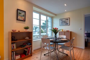 A dining area with a round table, white chairs, and a bookshelf with books and decorative items. - Dolmen Horizon - A deux pas du front de mer (Carnac)