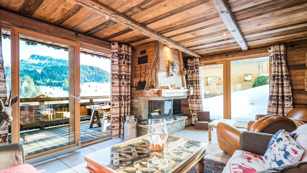 Ein gemütliches Holzhaus-Interieur mit Kamin, einem Glastisch und Blick auf die verschneite Landschaft.