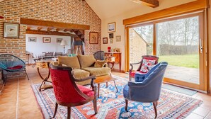 A spacious living room with a brick wall, wooden beams, a large sliding glass door, and a mix of modern and classic furniture. - Coeur de Sologne - Proximité Châteaux de la Loire (Saint-Viâtre)