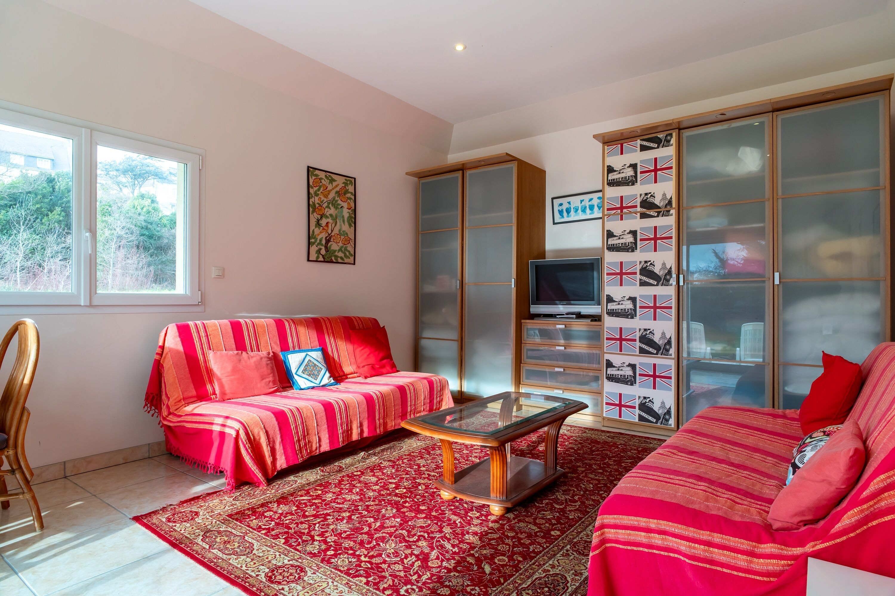 Un salon avec un canapé rouge, une table basse en verre et un téléviseur posé sur un meuble. Il y a une grande fenêtre et une bibliothèque remplie de photos.