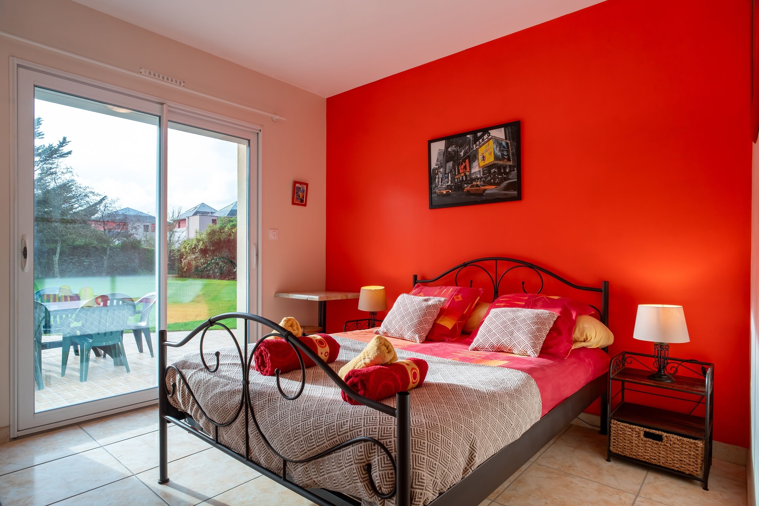 Ein Schlafzimmer mit roter Wand, einem Bett mit gemusterten Bettbezügen, zwei Nachttischen mit Lampen und Blick durch eine Schiebetür auf einen Außenbereich mit Tisch und Stühlen.