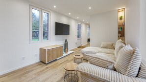 A modern living room with a sofa set, a wooden cabinet, a flat-screen TV mounted on the wall, and a bookshelf. - Aux Magnolias - Maison pour 6p (Silly-Tillard)