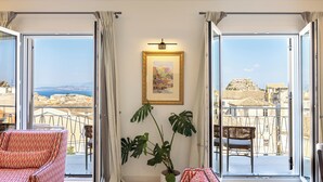 En rummelig balkon med udsigt over en kystby, møbleret med en rød sofa med mønster og en fletstol, samt en potteplante.