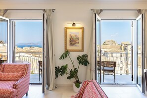 A spacious balcony with a view of a coastal cityscape, furnished with a red patterned sofa and a wicker chair, and a potted plant. - Terra Di Corfu, Αpartment (Kerkira)