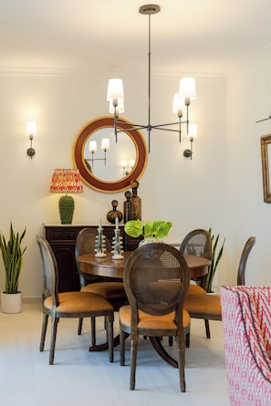 Ein Essbereich mit einem runden Tisch, sechs Stühlen, einer Chandelier, Wandleuchten, einem Spiegel und Dekorationselementen.