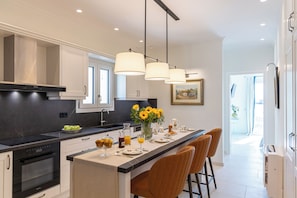A modern kitchen with a central island, overhead pendant lighting, and a dining area with bar stools. - Terra Di Corfu - Panoramic Penthouse, Αpartment (Kerkira)
