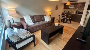 Una sala moderna con un sofá marrón, dos sillones, una mesita de centro y una zona de comedor con sillas y una mesa. Se observa una cocina al fondo con un microondas y una cafetera.