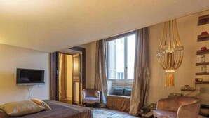 Habitació d'hotel amb un llit, una televisió, una cadira, un sofà, una finestra amb cortines i un penjoll.
