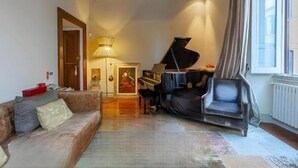 Un saló amb un piano, un sofà i una cadira. Hi ha una porta que dona a una altra habitació i una finestra amb cortines.