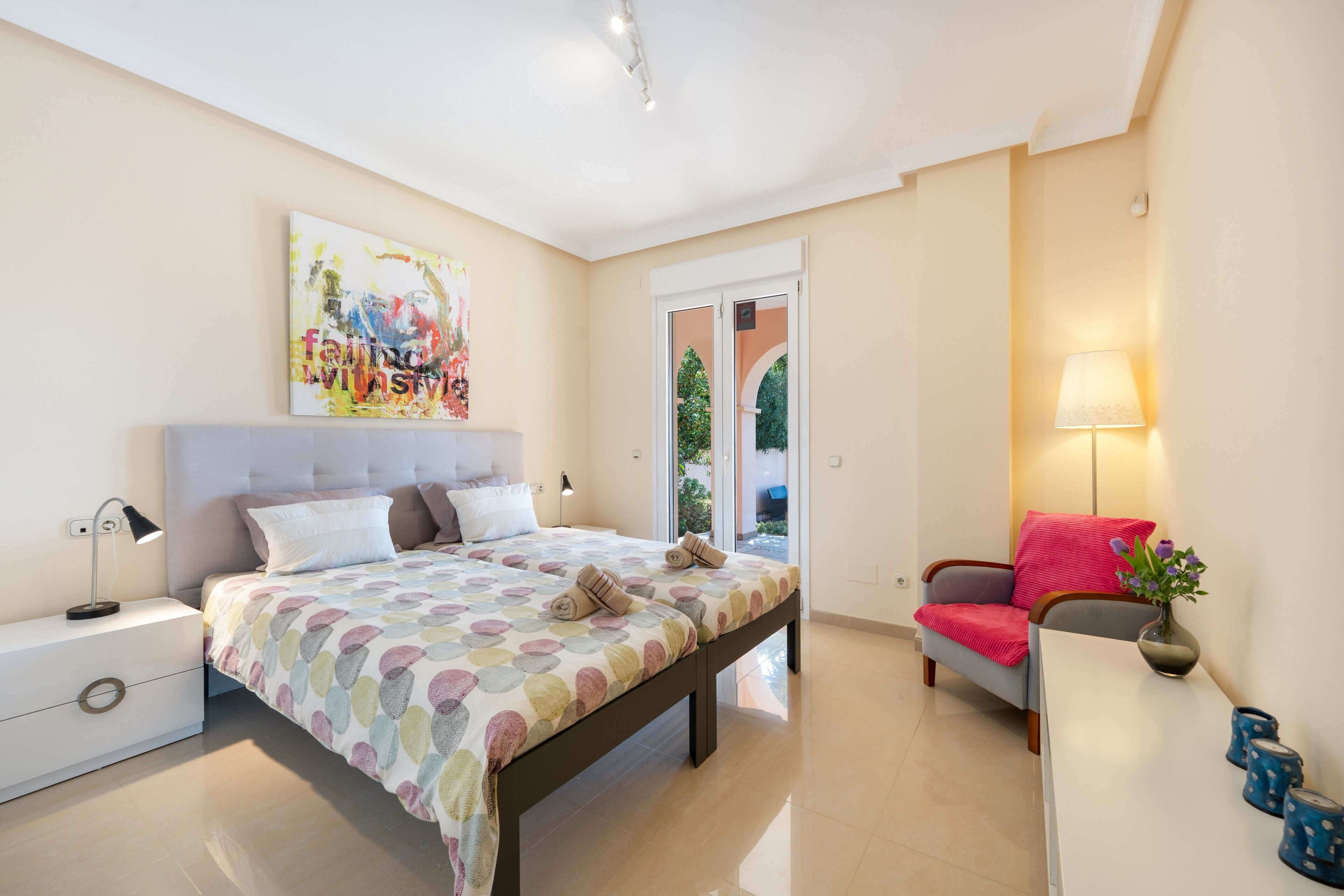 Un dormitorio con cama, mesita de noche, lámpara, silla y un cuadro en la pared.