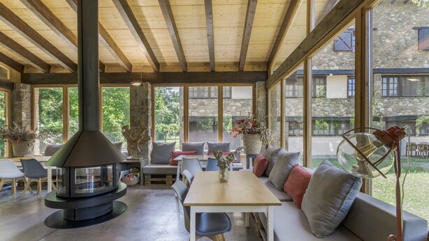A covered outdoor area with a stone wall, wooden beams, a fireplace, seating, and large windows.