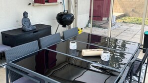 Ulkoterassi, jossa on lasipöytä, tuoleja, Buddha-patsas, grilli ja punainen ovi.