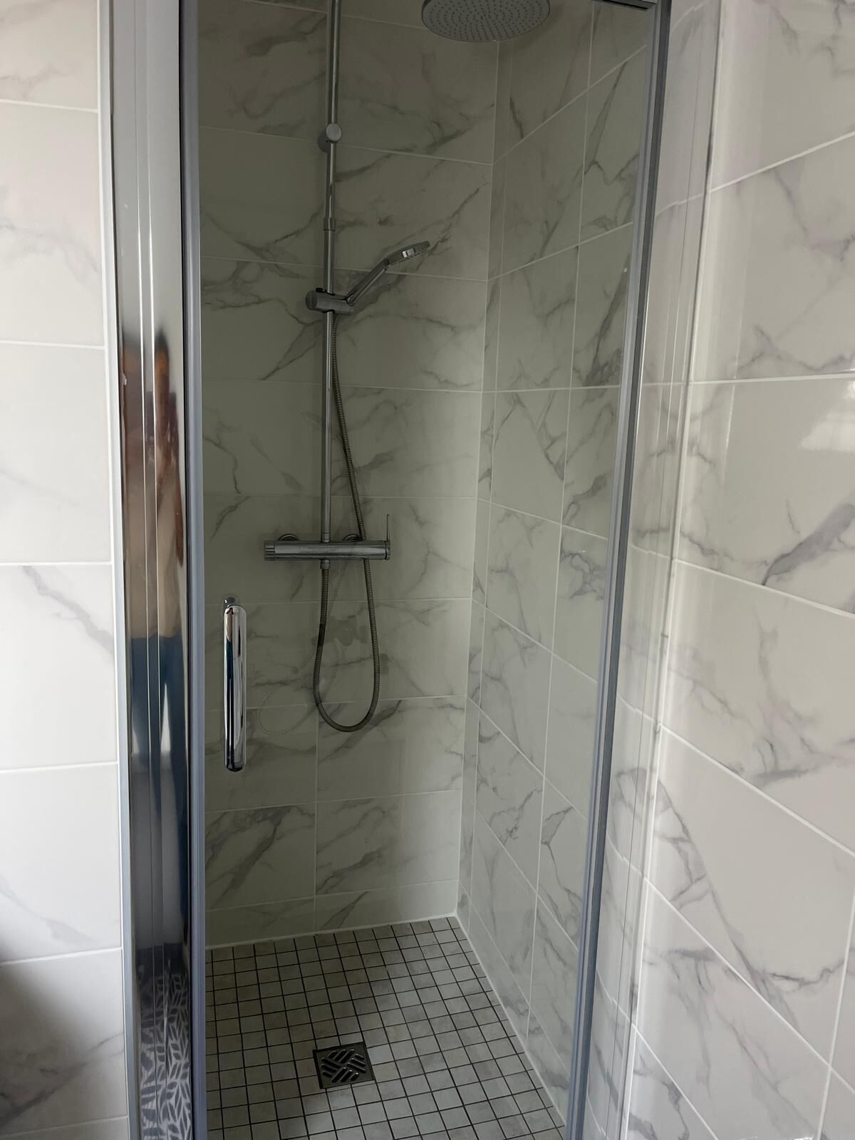 Eine begehbare Dusche mit Glastrennwand, Handbrause und Deckenbrause, Wände mit Marmoroptik und ein Fliesenboden.