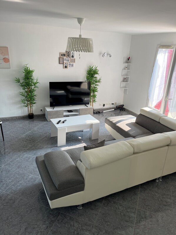 A modern living room with a large flat-screen TV, a white sofa, a coffee table, and two potted plants. - Le clos de Précy- House 132 m² 10 pers Private garden Quiet 20 min Disneyland (Précy-sur-Marne)