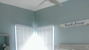 Une chambre à coucher comprenant un lit, une commode, un ventilateur de plafond et une fenêtre avec des stores.