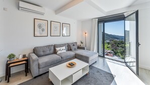 A modern living room with a grey sectional sofa, a white coffee table, and a large sliding glass door leading to a balcony with a view of a hilly landscape.