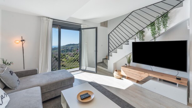 A modern living room with a sofa, a coffee table, a TV, a staircase, and a balcony with a view.