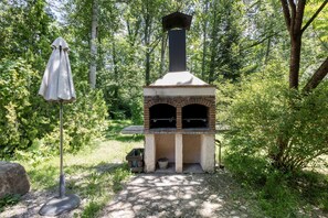 A brick outdoor pizza oven with a chimney, surrounded by trees and a picnic table.