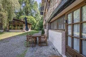 A patio with a wooden table and chairs, a covered umbrella, and a stone building with large windows.