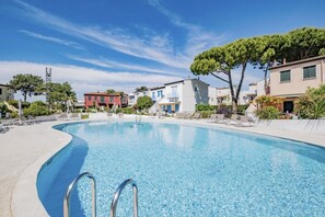 A swimming pool with a curved edge, surrounded by lounge chairs and a row of buildings with balconies. - Villaggio dei Gabbiani - Carraro Immobiliare Jesolo (Lido di Jesolo)