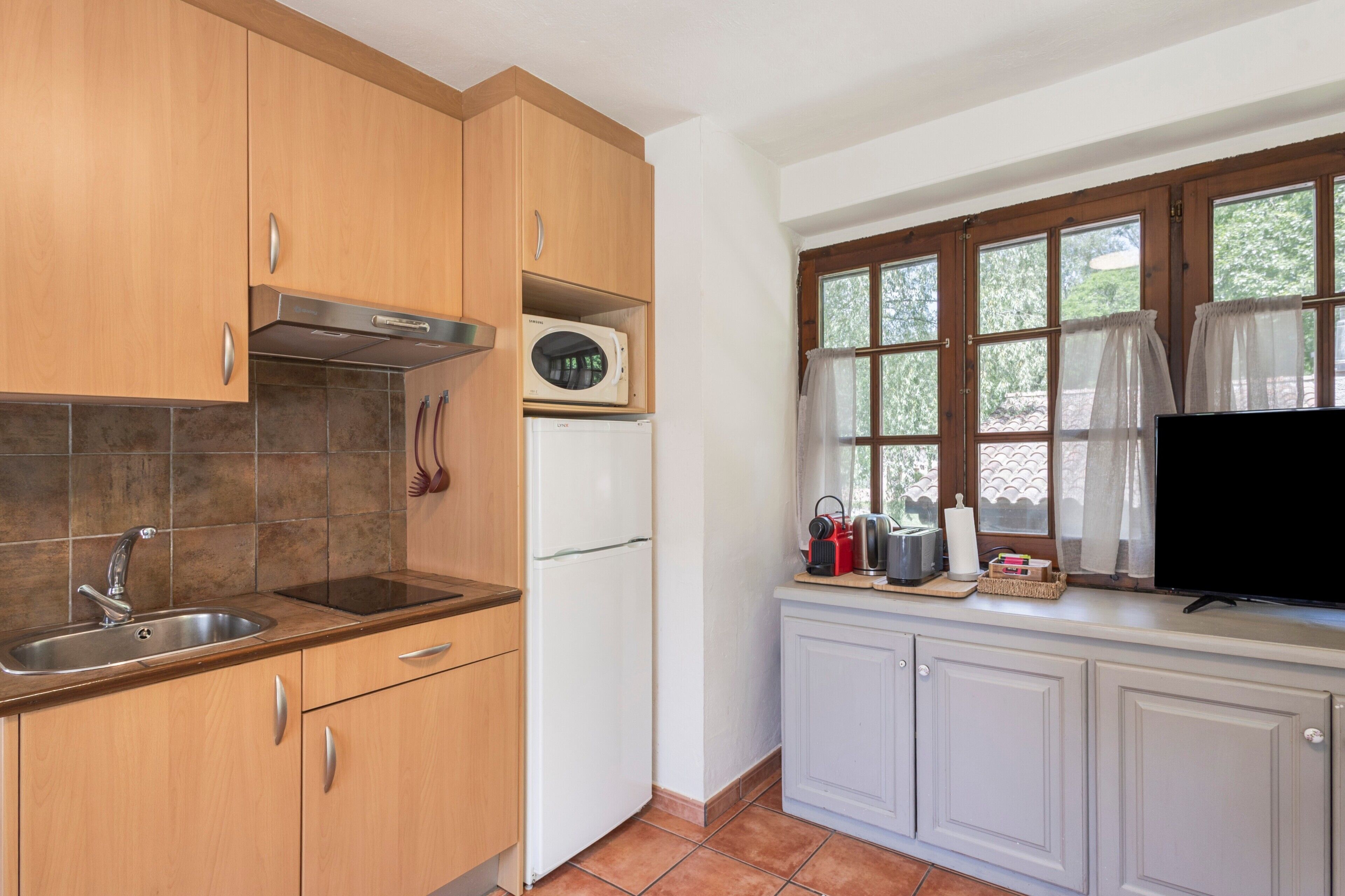 A kitchen with wooden cabinets, a stainless steel sink, a microwave, a refrigerator, and a window with curtains.