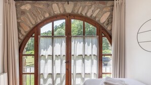 Una habitación con ventanal de arco de piedra, cama con ropa blanca y vistas a la vegetación.
