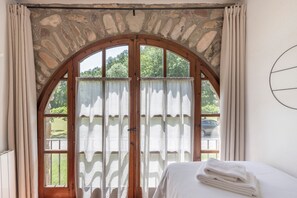 Een kamer met een gewelfraam venster, een bed met witte lakens, en uitzicht op groen.