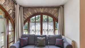 Una habitación con pared de piedra, un amplio ventanal arqueado y un sofá gris con cojines.