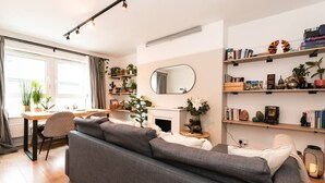 A modern living room with a grey sofa, a dining table with chairs, a fireplace, and shelves with books and decorative items. - Modern & Convenient 2-Bed Flat Near Tube – Heart of London (London)
