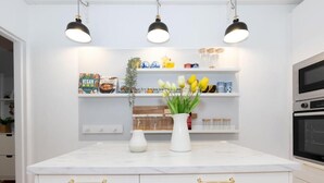 A modern kitchen with white cabinetry, gold handles, and a marble countertop. There are three pendant lights above the counter, and a vase with yellow flowers on the island. Shelves above the counter hold various kitchen items and decorative objects. - Modern & Convenient 2-Bed Flat Near Tube – Heart of London (London)
