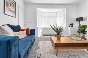 Un salon moderne avec un canapé bleu, une table basse en bois et une grande fenêtre dotée de stores blancs.