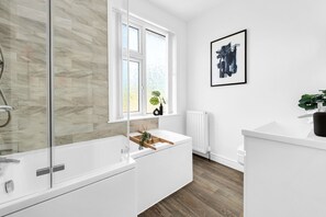 A modern bathroom with a glass shower enclosure, a white bathtub, a window with frosted glass, a radiator, and a framed abstract painting on the wall. - 2BDR Stanley House | Free Parking | Private Garden (Stoke-on-Trent)
