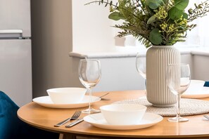 A dining table with white plates, bowls, and wine glasses, accompanied by a vase of green foliage. - 2BDR Stanley House | Free Parking | Private Garden (Stoke-on-Trent)