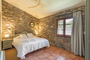 A bedroom with a stone wall, a bed with white linens, a ceiling fan, a window with curtains, and a nightstand with a lamp. - El Roure I de Can Torrotes (Sant Martí del Clot)