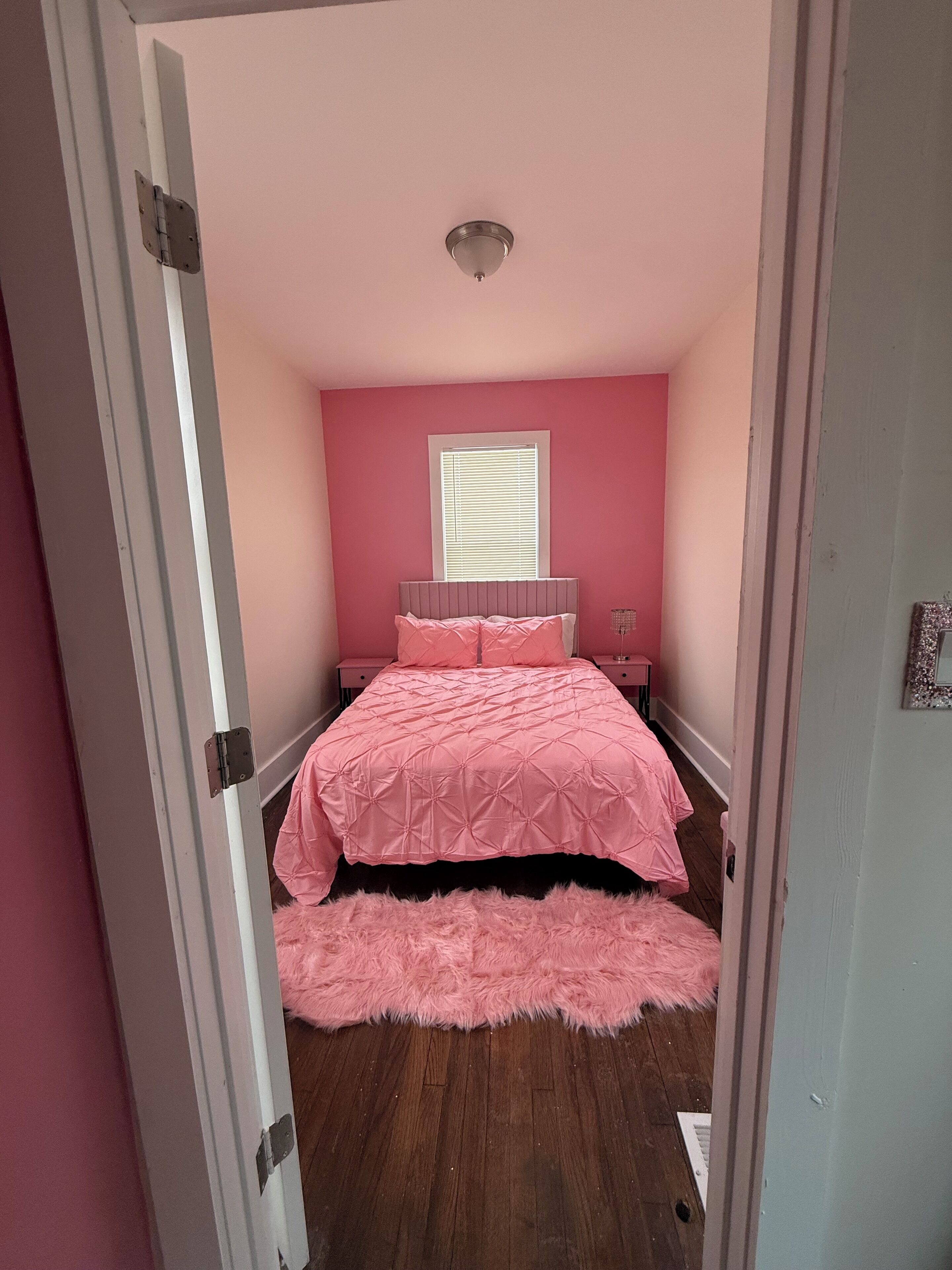 Ein Schlafzimmer mit pinkfarbener Wand, einem Bett mit pinkfarbener Bettdecke, einem flauschigen pinkfarbenen Teppich und einem Fenster mit Jalousien.