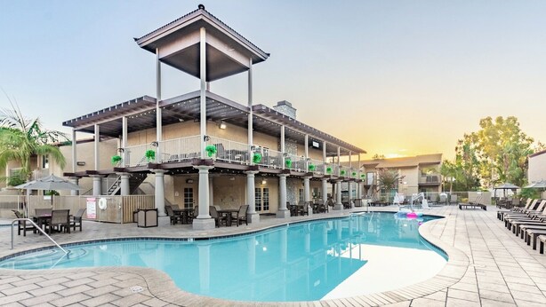 Un área de piscina con zona de estar cubierta, sillones reclinables y un edificio con varios balcones.